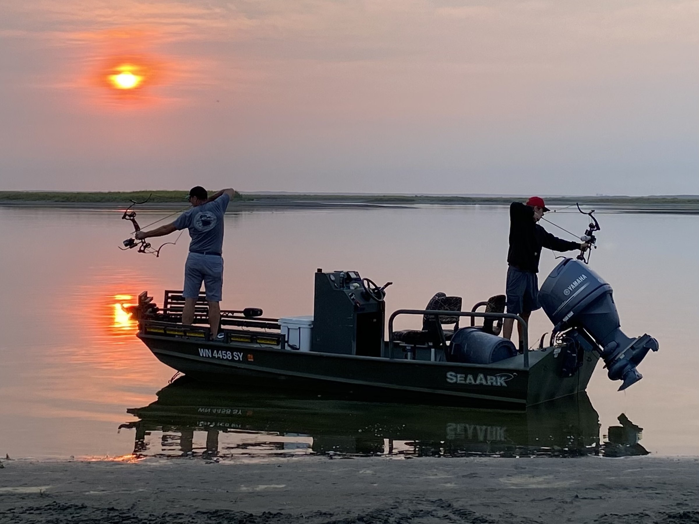 Two bowfishermen at full draw on a SeaArk boat at sunrise